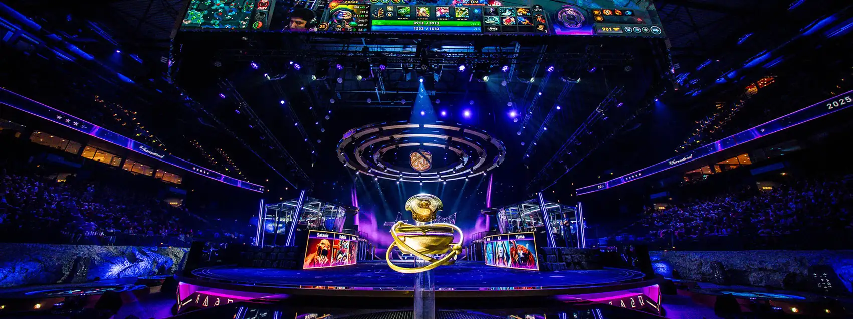 Grote esports-arena met een Dota 2-toernooipodium, een kampioenschapstrofee in het midden en enorme schermen waarop live gameplay wordt getoond.